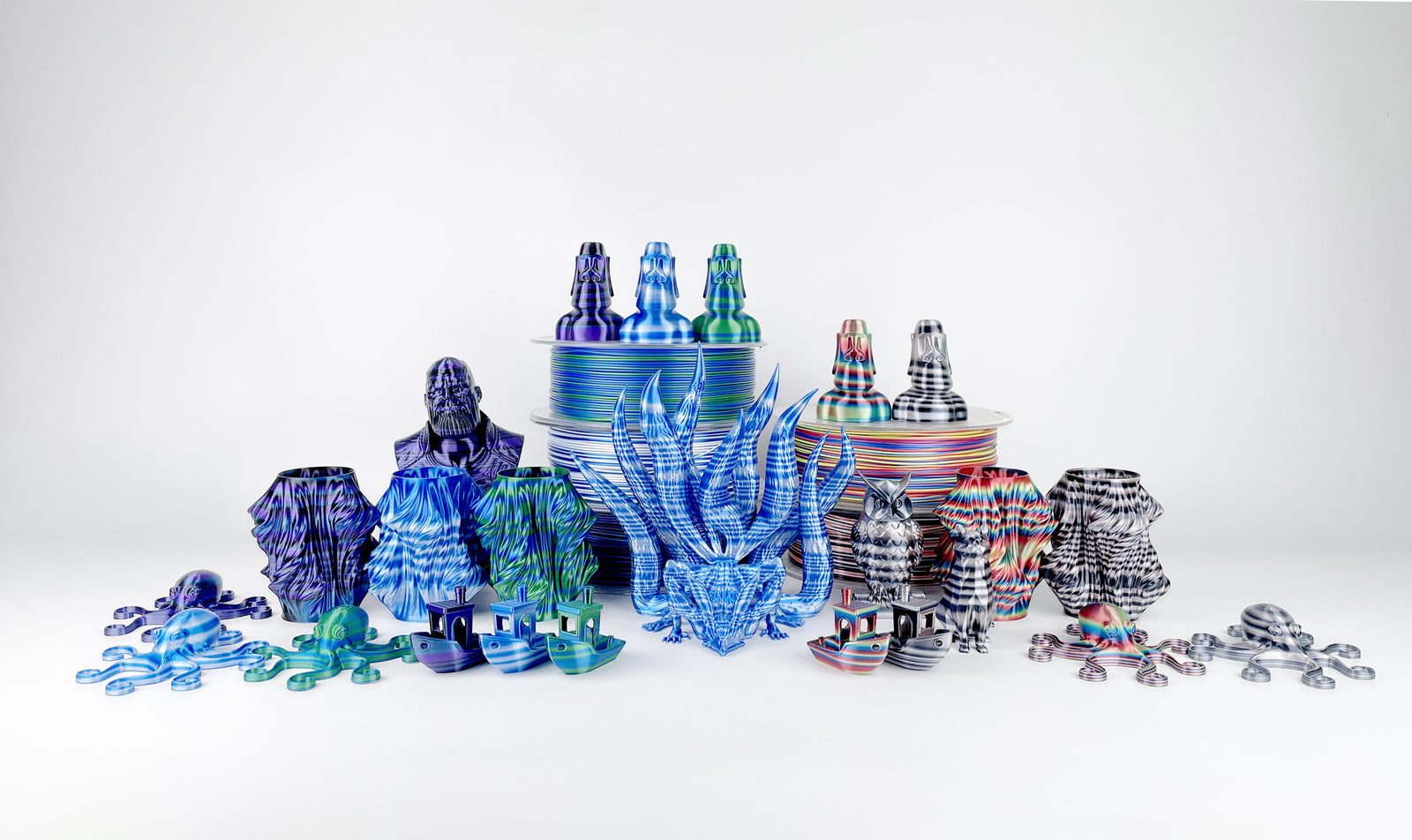EIBOS Artism Series II Ribbons PLA filament spools with colourful striped 3D prints, including a dragon, Moai statues, vases, owls, Benchy boats and octopuses.
